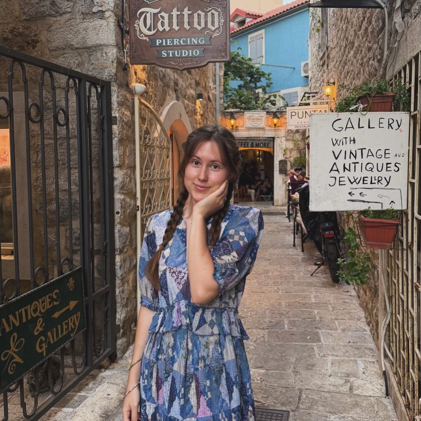 Anastasiia in a blue patterned dress on a narrow stone street lined with small shops